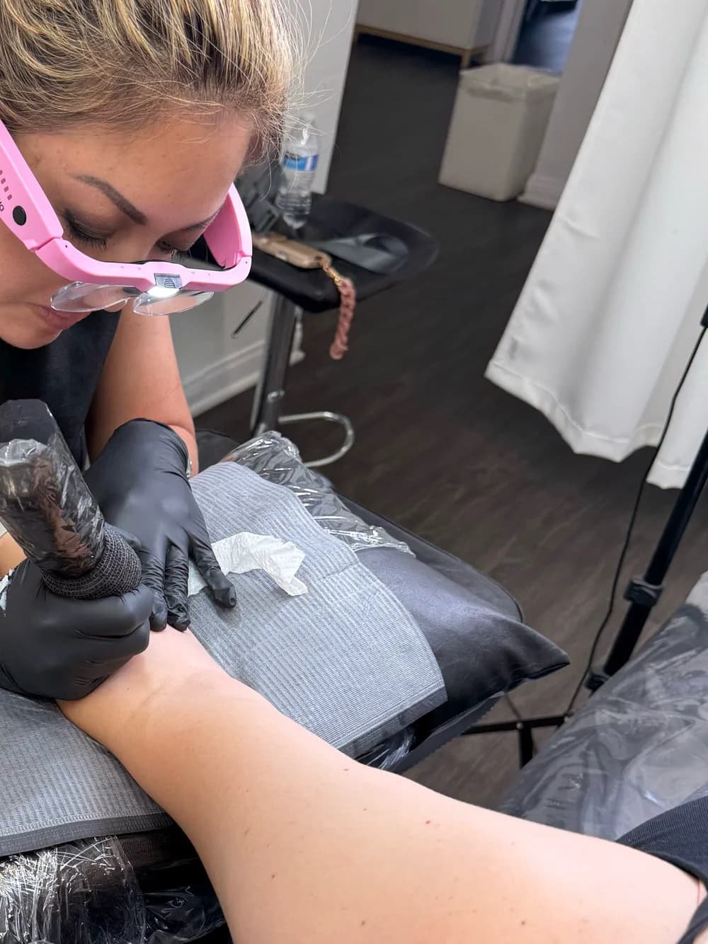 Cindy Keyes wearing magnifying glasses while creating a fine-line tattoo on a client’s forearm.